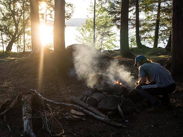 Student starts a fire at a campsite while the sun sets over a lake in the background.