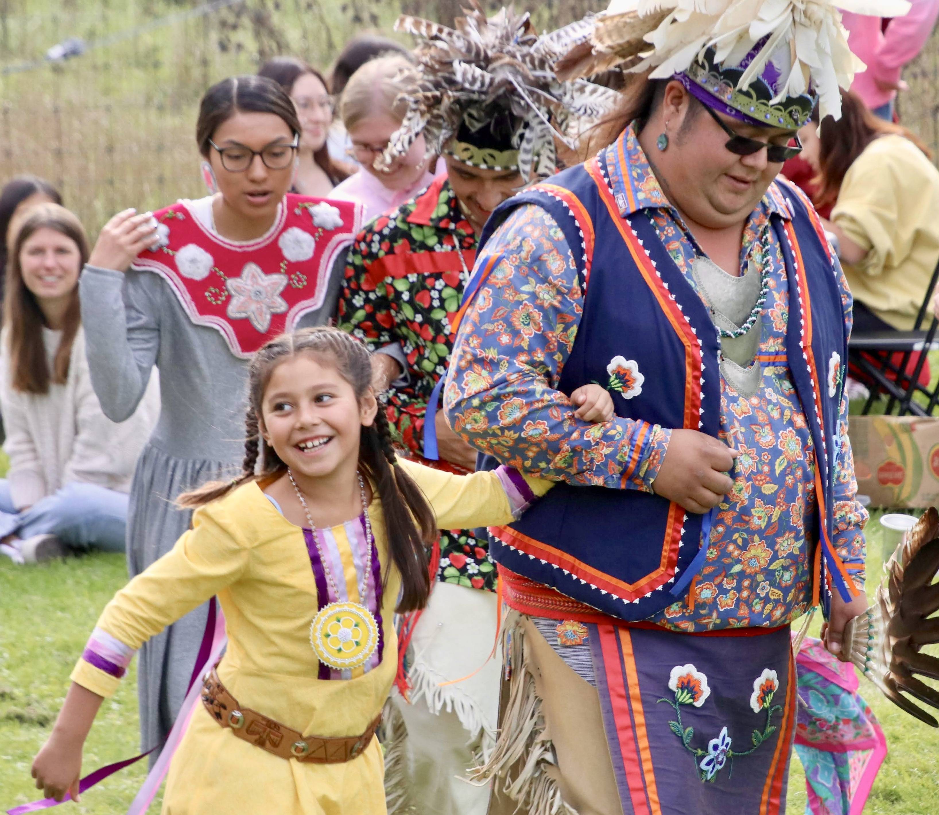 A girl in a yellow dress links arms with one of the Haudenosaunee dancers at the Fall Festival.