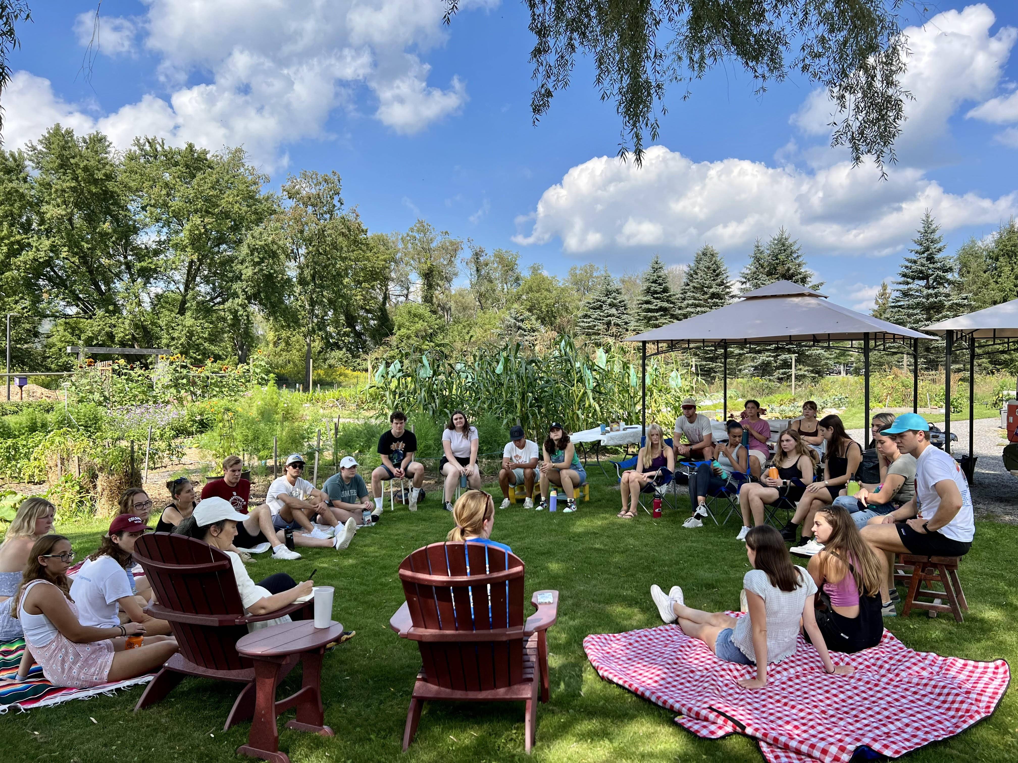 A group of students sit on the ground in a circle at the Community Garden. The lush, green garden stands out under the brilliant blue sky.