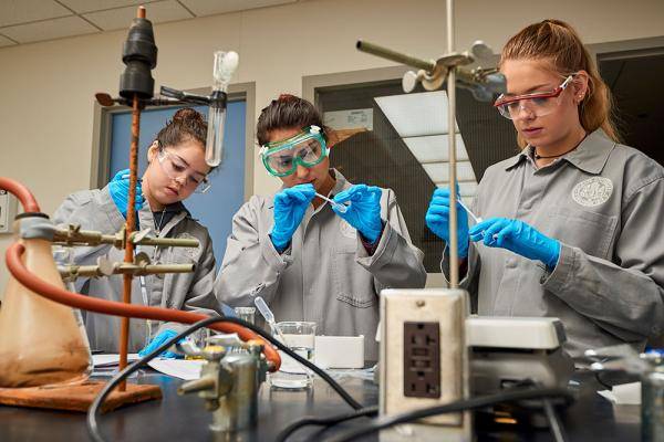 Three women, wearing gloves and safety glasses, work together in lab