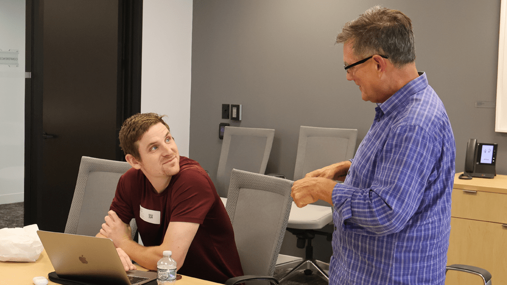 Mentor Andres Echenique, standing, speaks with an alum entrepreneur, seated.