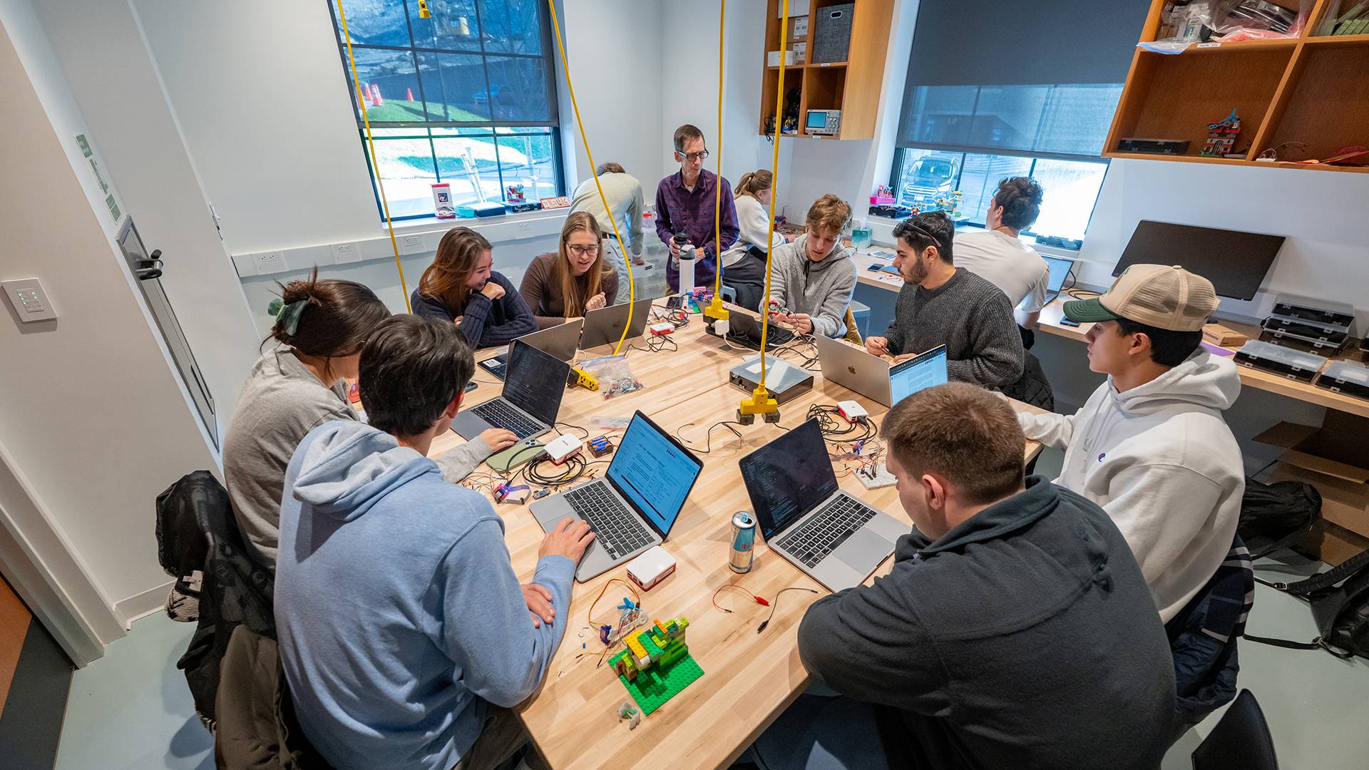 Computer science students sit at a square table, each with a laptop in front of them. A professor stands in the background.