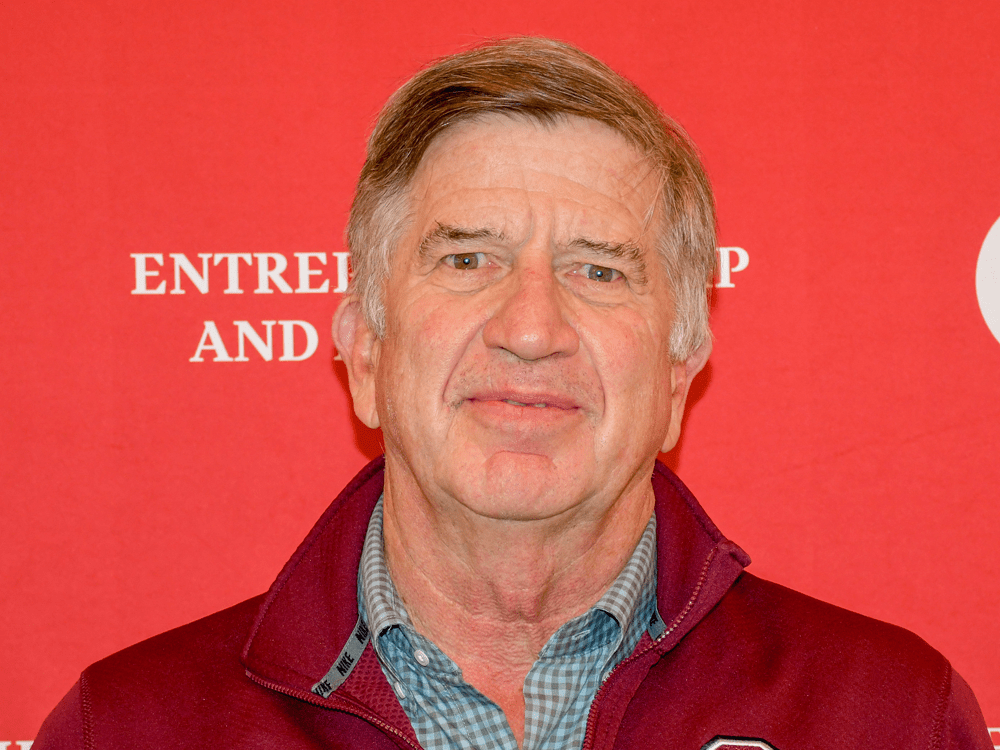 A man with light brown hair and a slight smile poses for a headshot in front of a bright red backdrop featuring white "C" logos and the words "Entrepreneurship and Innovation." He wears a maroon zip-up jacket over a blue checkered shirt.