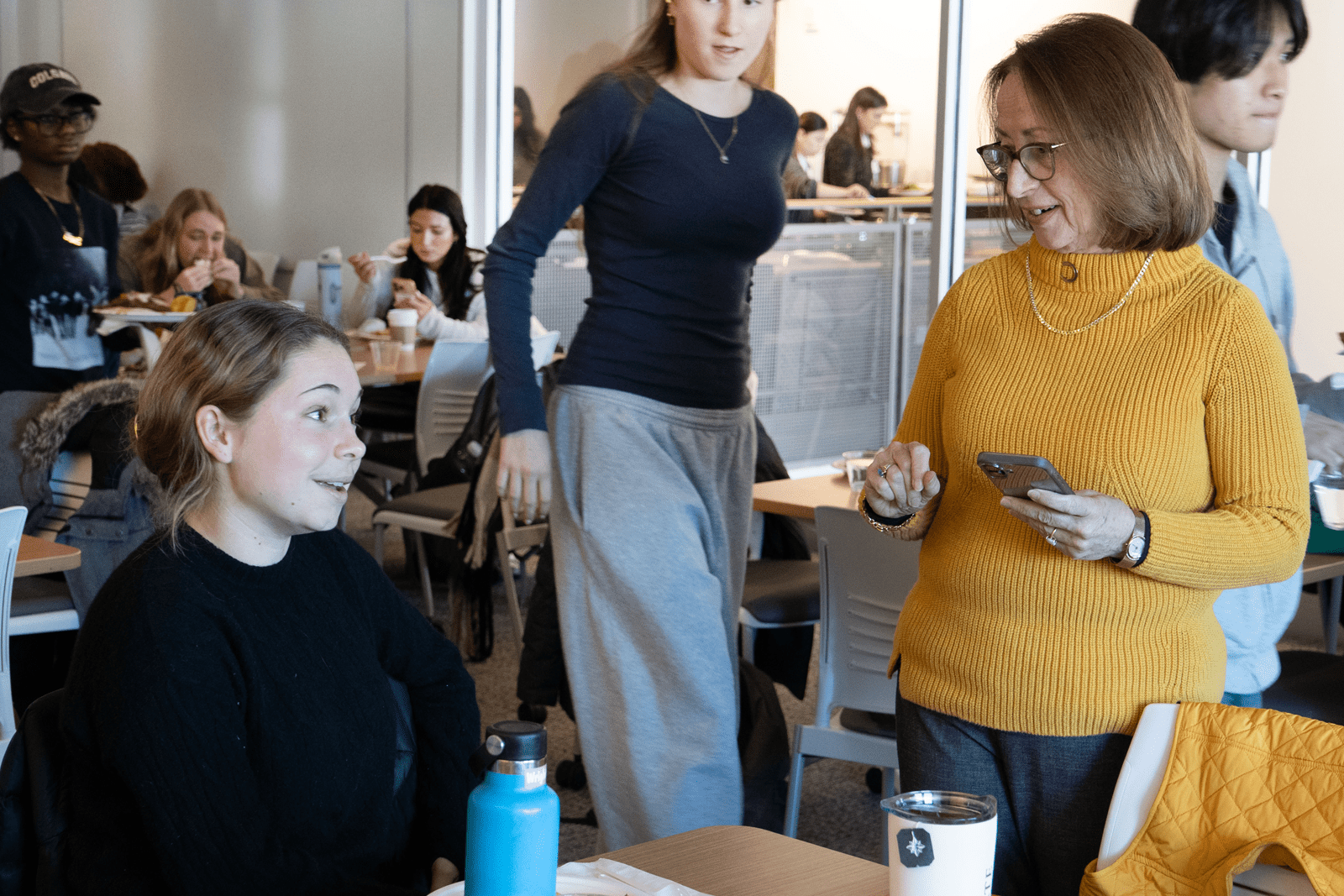 In a brightly lit indoor setting, a woman in a yellow sweater stands and holds a smartphone while engaging in a friendly conversation with a seated young woman in a black sweater. The background shows a bustling common area with several other students eating, talking, and walking through the space.