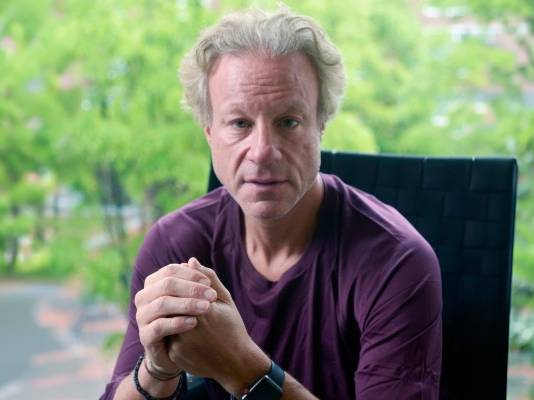 A medium close-up shows a man with light, wavy hair and a neutral expression sitting in a black woven chair with his hands clasped in front of him. He is wearing a dark purple shirt and a smartwatch, positioned against a background of bright green trees visible through a window.