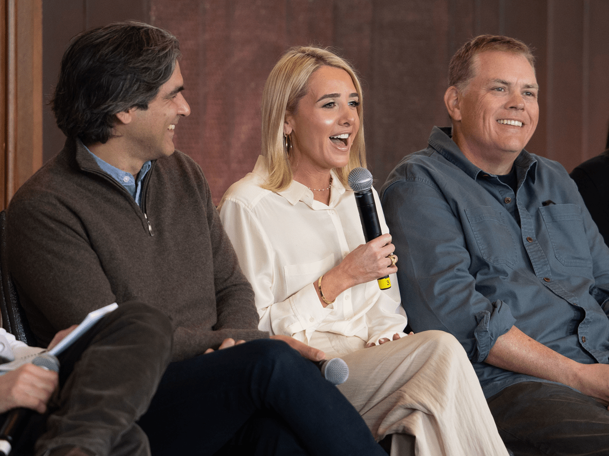 Three people sit side-by-side on a panel, with the woman in the center speaking into a microphone while the men on either side of her listen and smile. She wears a cream-colored blouse and tan trousers, while the men are dressed casually in a brown quarter-zip sweater and a grey button-down shirt.