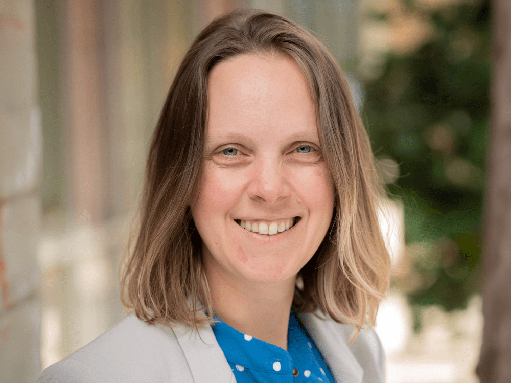 A headshot of a smiling woman with light brown, shoulder-length hair and blue eyes, wearing a light gray blazer over a blue polka-dot shirt. She is positioned outdoors with a soft-focus background of greenery and architectural elements, creating a bright and professional atmosphere.