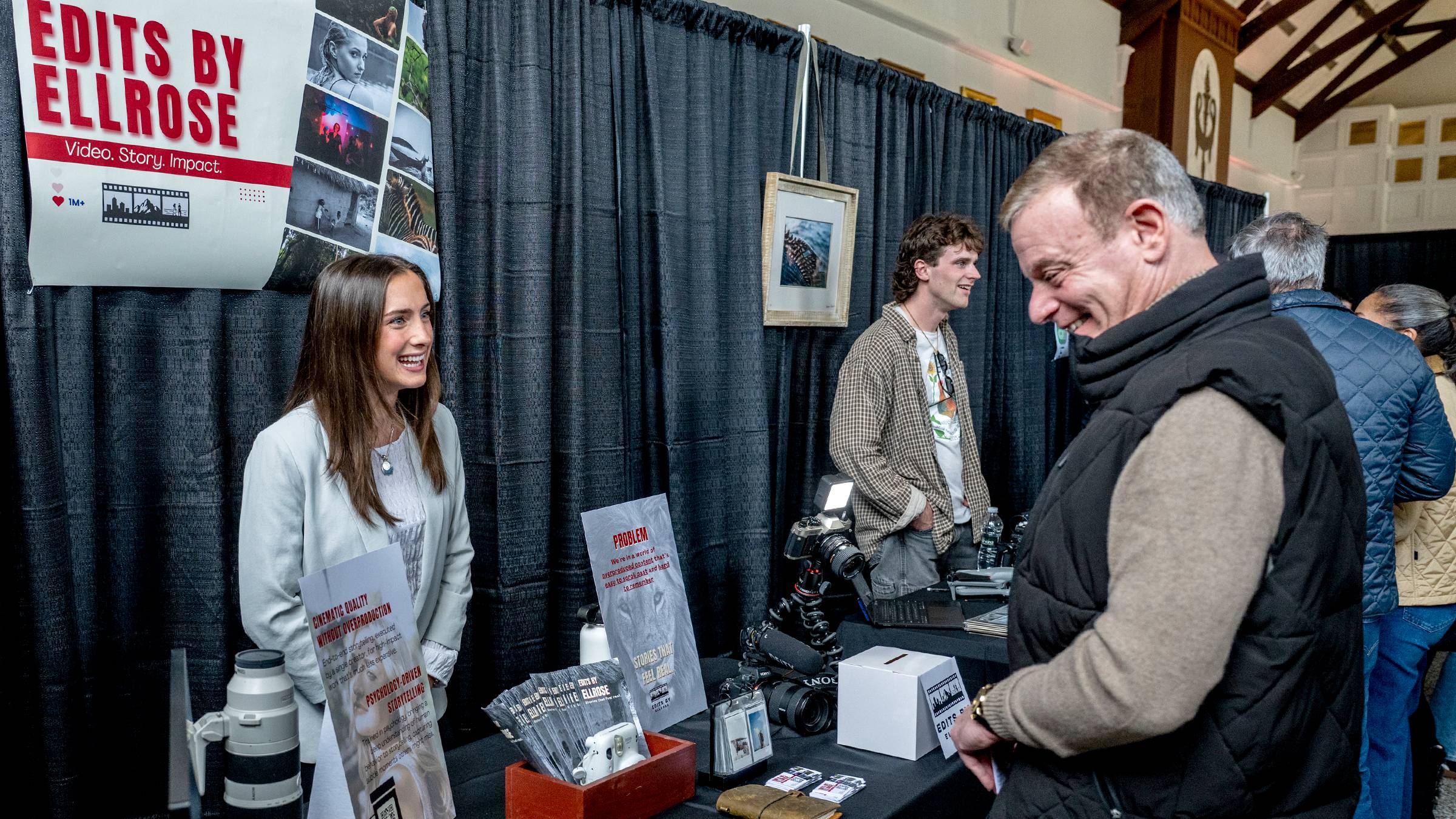 A young woman smiles while engaging with a man at her promotional booth for "Edits by Ellrose," which features a large banner displaying her video and storytelling services. The booth is professional and detailed, showcasing cameras, photography equipment, and informational signage against a backdrop of black curtains at a crowded event.