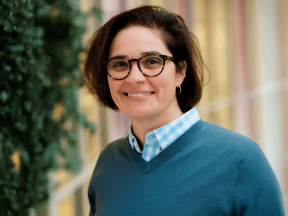 A professional headshot of a woman with short dark hair and glasses, wearing a teal v-neck sweater over a blue and white checkered shirt. She is smiling warmly against a soft-focus background of greenery and light-colored architectural columns.