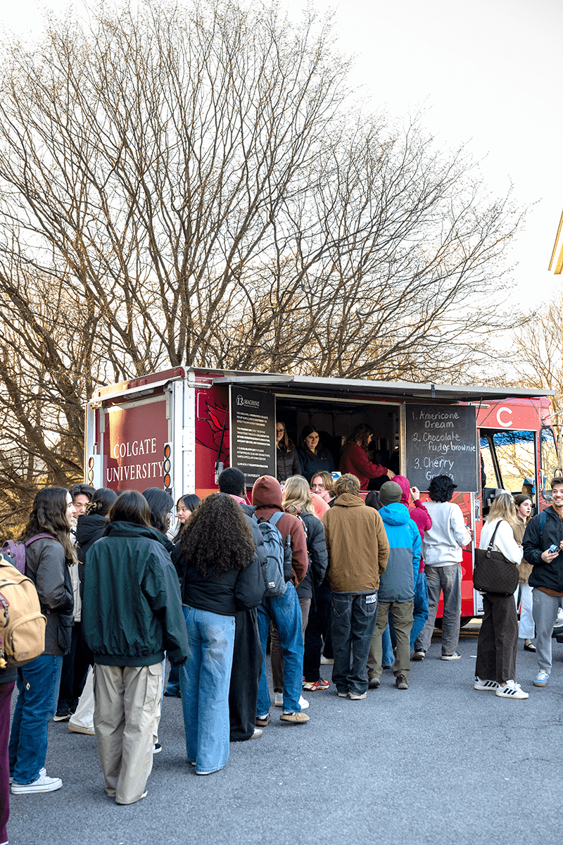 A large crowd of students stands in a long line outdoors to order from a maroon Colgate University food truck parked beneath bare, wintry trees. The truck's serving window features a chalkboard menu listing Ben & Jerry's ice cream flavors, including American Dream and Chocolate Fudge Brownie.