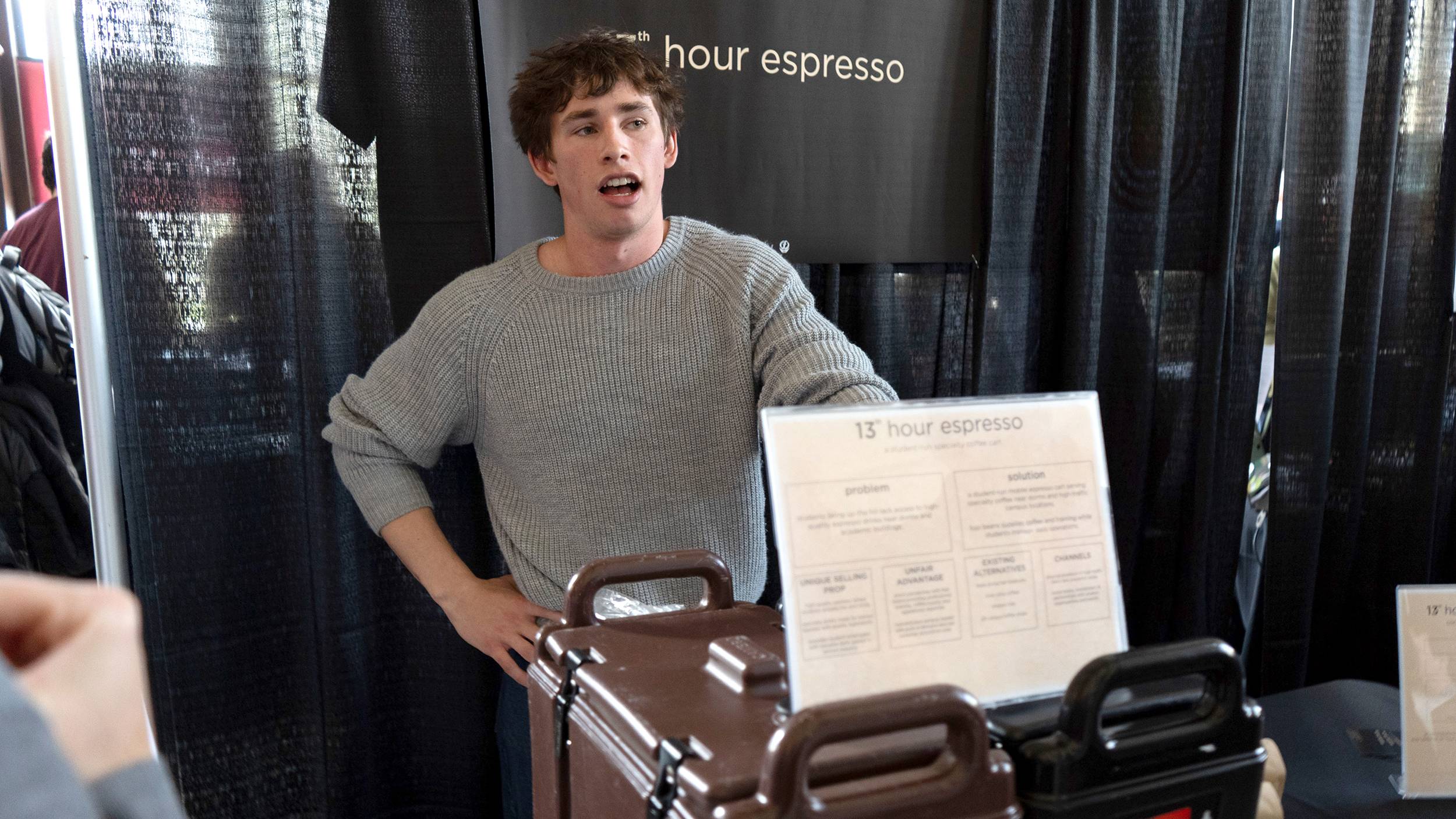 A young man with messy brown hair and a grey knit sweater stands mid-speech in front of a dark booth backdrop featuring a sign that reads "13th hour espresso." In the foreground, a partially visible tabletop sign also displays the "13th hour espresso" branding alongside text sections labeled "problem" and "solution."