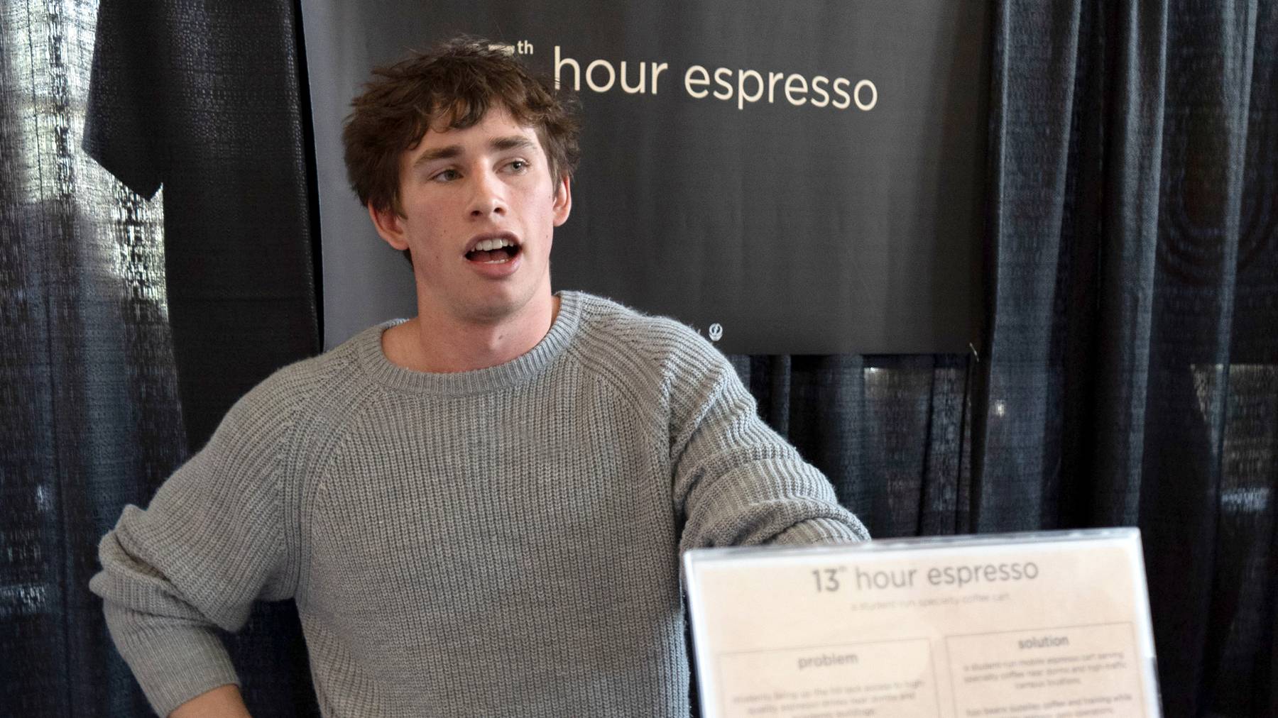 A young man with messy brown hair and a grey knit sweater stands mid-speech in front of a dark booth backdrop featuring a sign that reads "13th hour espresso." In the foreground, a partially visible tabletop sign also displays the "13th hour espresso" branding alongside text sections labeled "problem" and "solution."