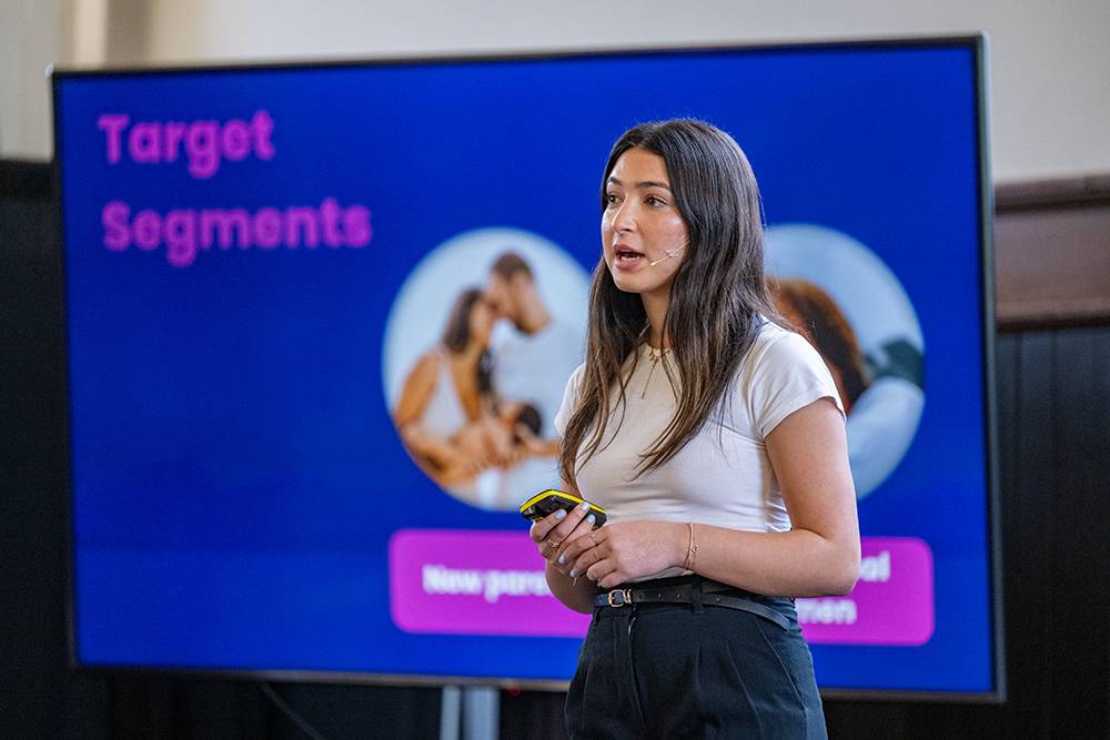 A student entrepreneur gives a presentation beside a screen reading “Target Segments,” holding a clicker while speaking in front of a slide with images.