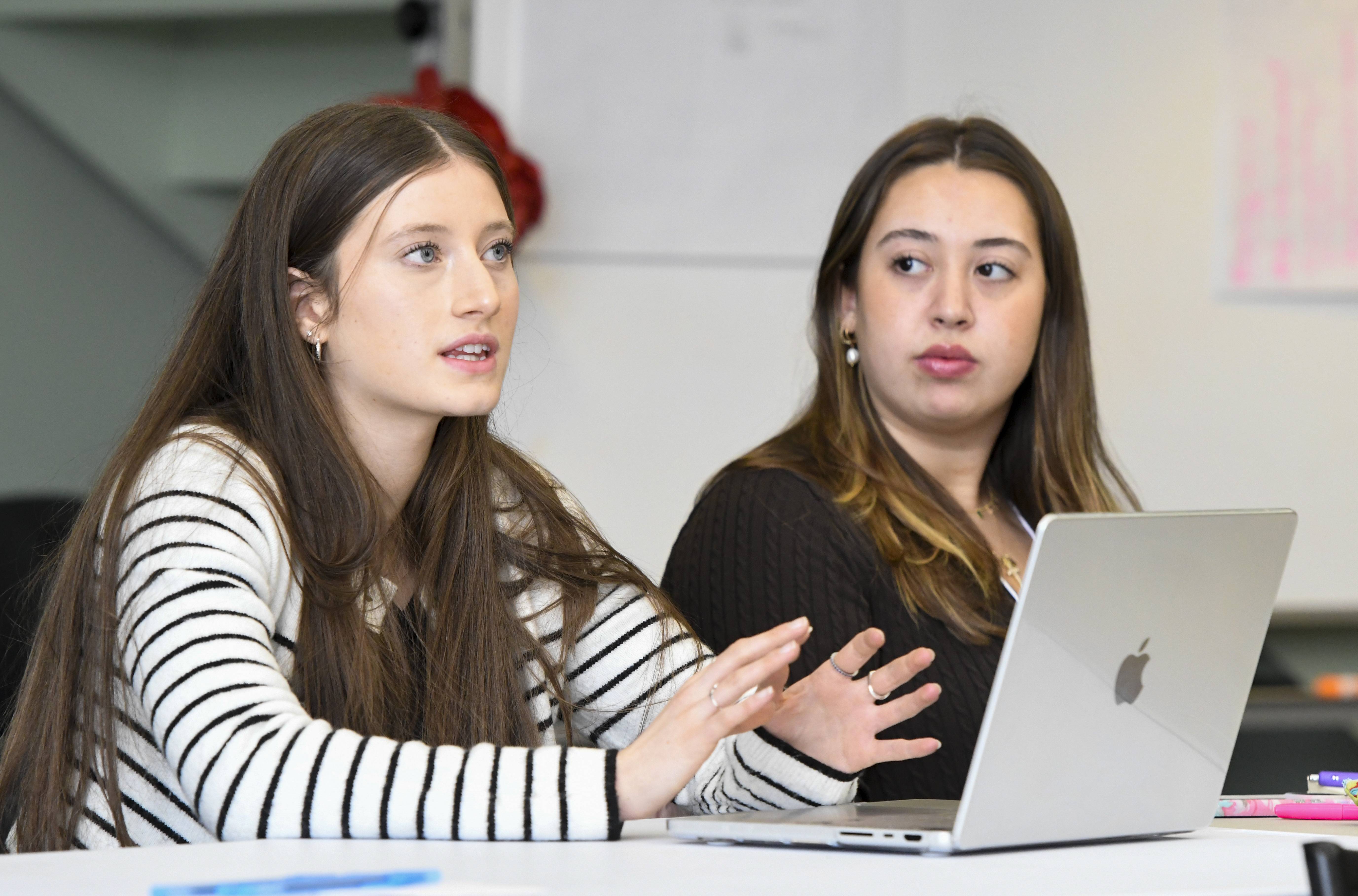Two students sit at a table during the summit, with one speaking and gesturing while the other listens beside an open laptop. 