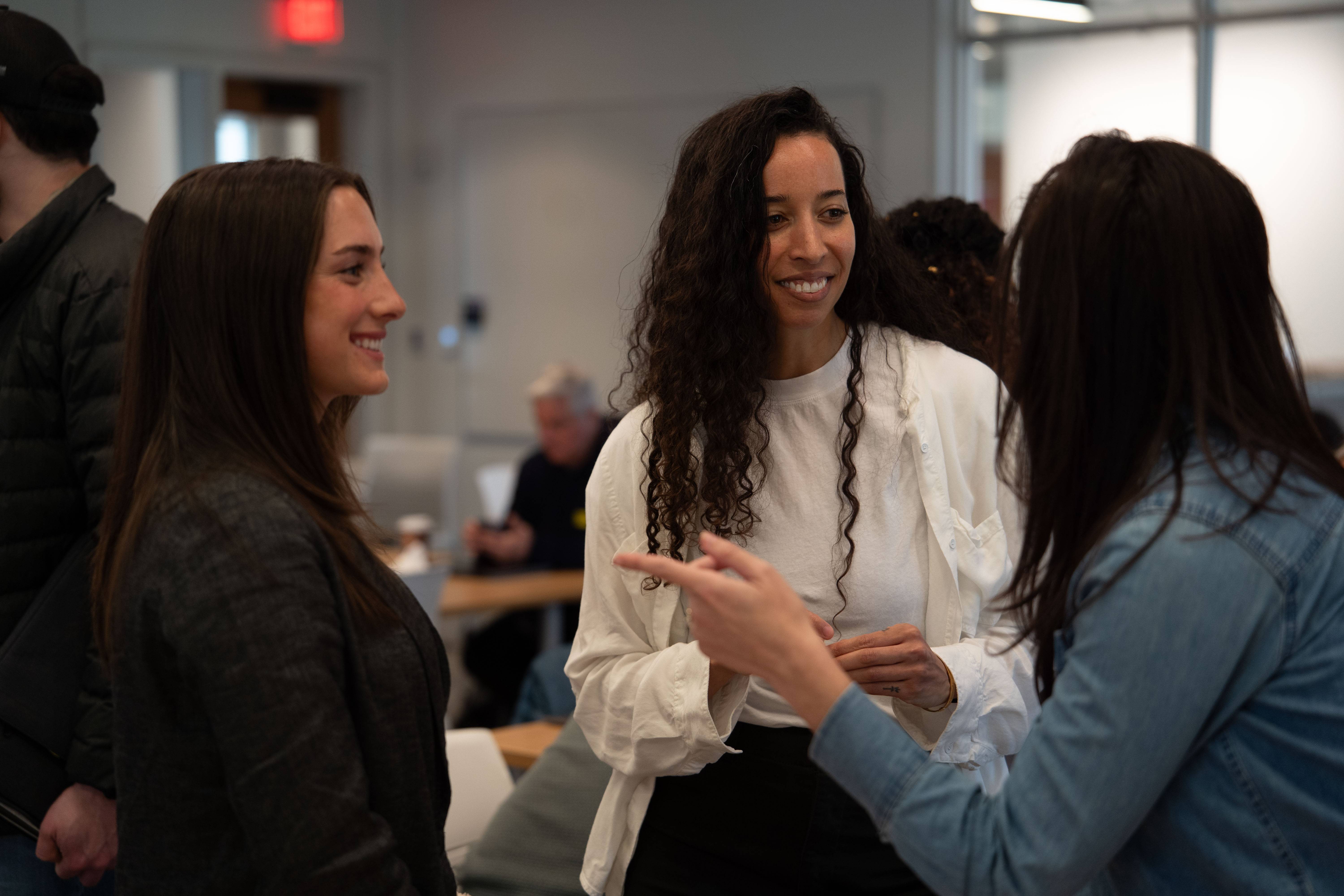 Three people stand in conversation indoors, smiling and gesturing as they talk in a bright gathering space.