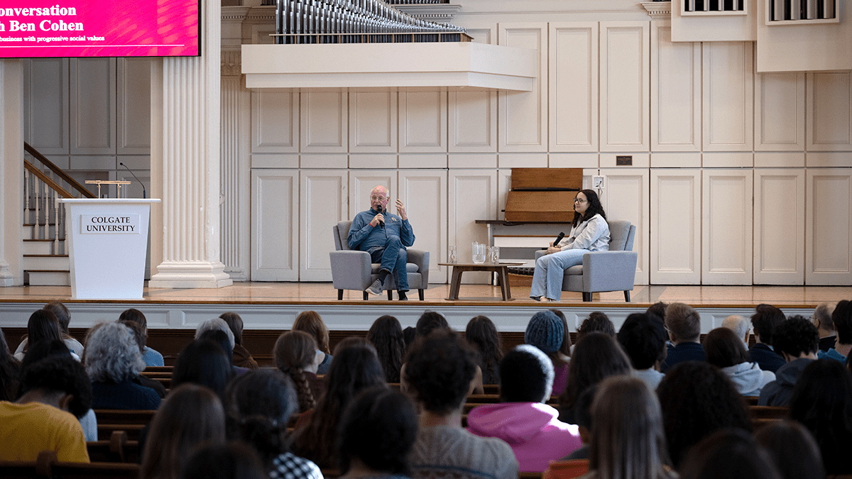 An older man with glasses and a younger woman with curly hair sit in grey armchairs facing each other during a discussion or interview. Both individuals hold microphones, and a small wooden coffee table with a water pitcher and glasses sits between them in a room with white architectural pillars.