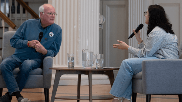An older man with glasses and a younger woman with curly hair sit in grey armchairs facing each other during a discussion or interview. Both individuals hold microphones, and a small wooden coffee table with a water pitcher and glasses sits between them in a room with white architectural pillars.
