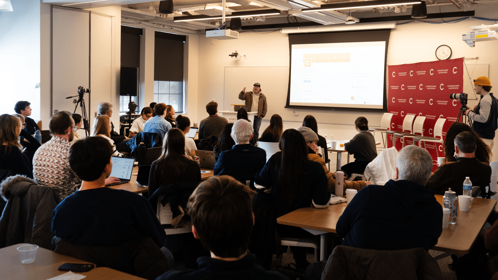 In a bright classroom setting, a man stands at the front of the room giving a presentation to a seated audience of students and adults. The presenter is positioned between a large projection screen displaying a website and a red backdrop labeled “ENTREPRENEURSHIP AND INNOVATION.”