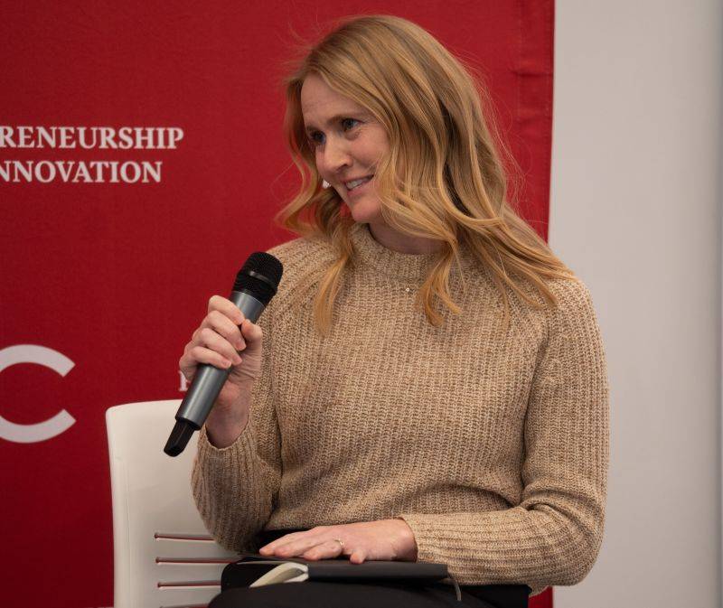 A woman with long blonde hair holds a microphone while seated in front of a red Entrepreneurship and Innovation backdrop.