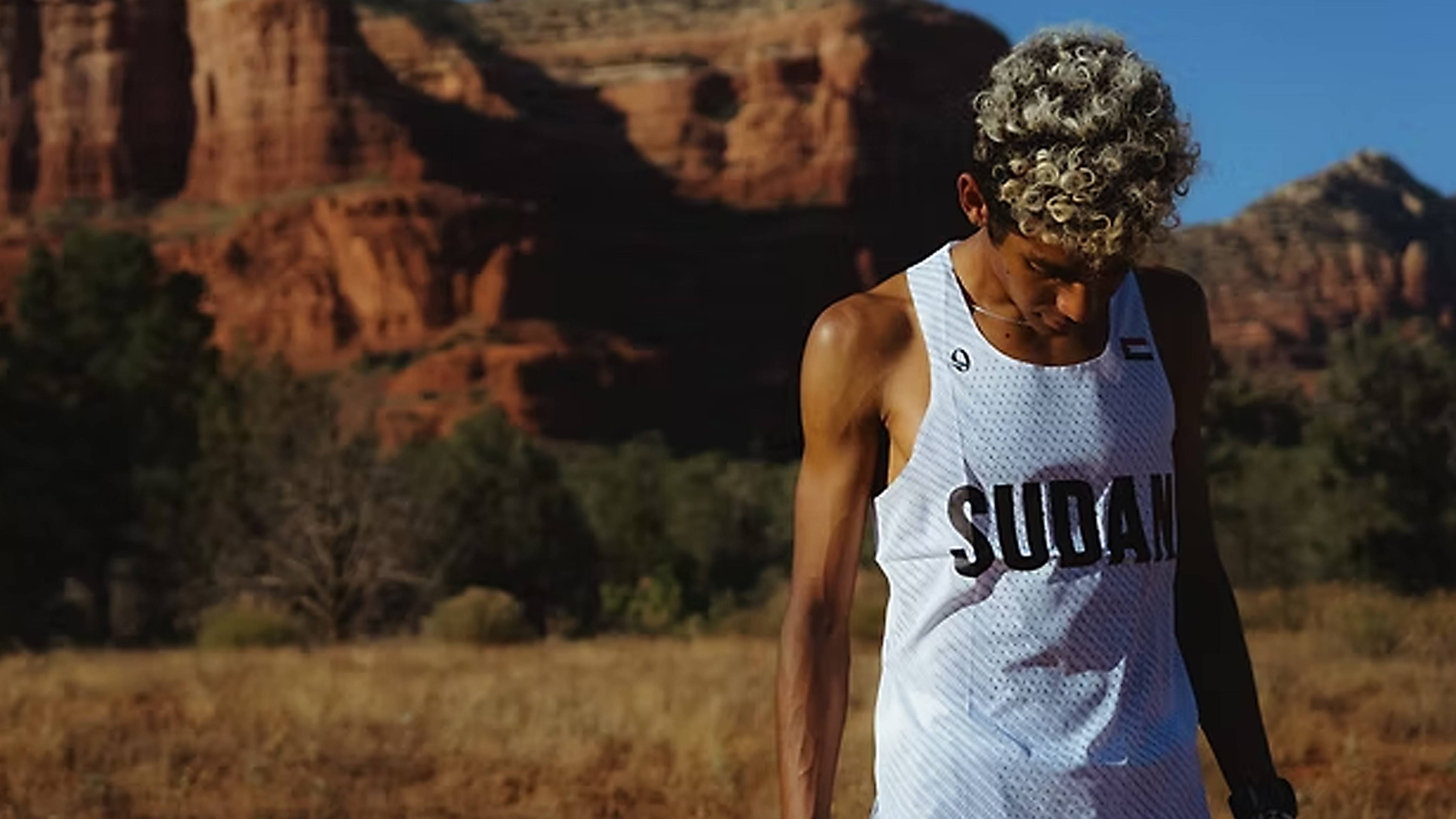 A runner with curly, frosted hair looks downward while standing in a dry, grassy field before a backdrop of massive red rock formations. He is wearing a white mesh tank top emblazoned with "SUDAN" across the front and a small flag on the left chest.