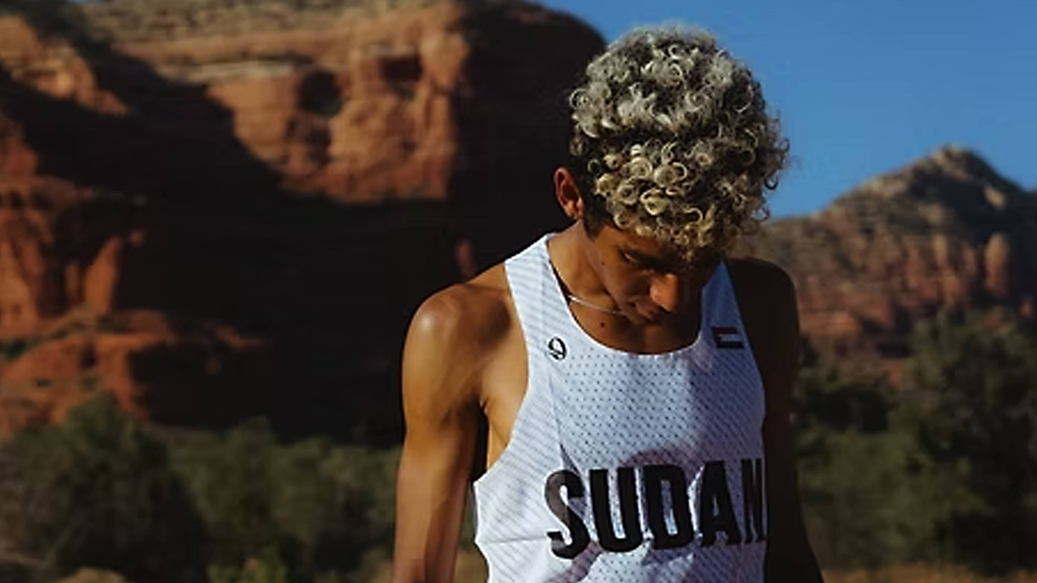 A runner with curly, frosted hair looks downward while standing in a dry, grassy field before a backdrop of massive red rock formations. He is wearing a white mesh tank top emblazoned with "SUDAN" across the front and a small flag on the left chest.