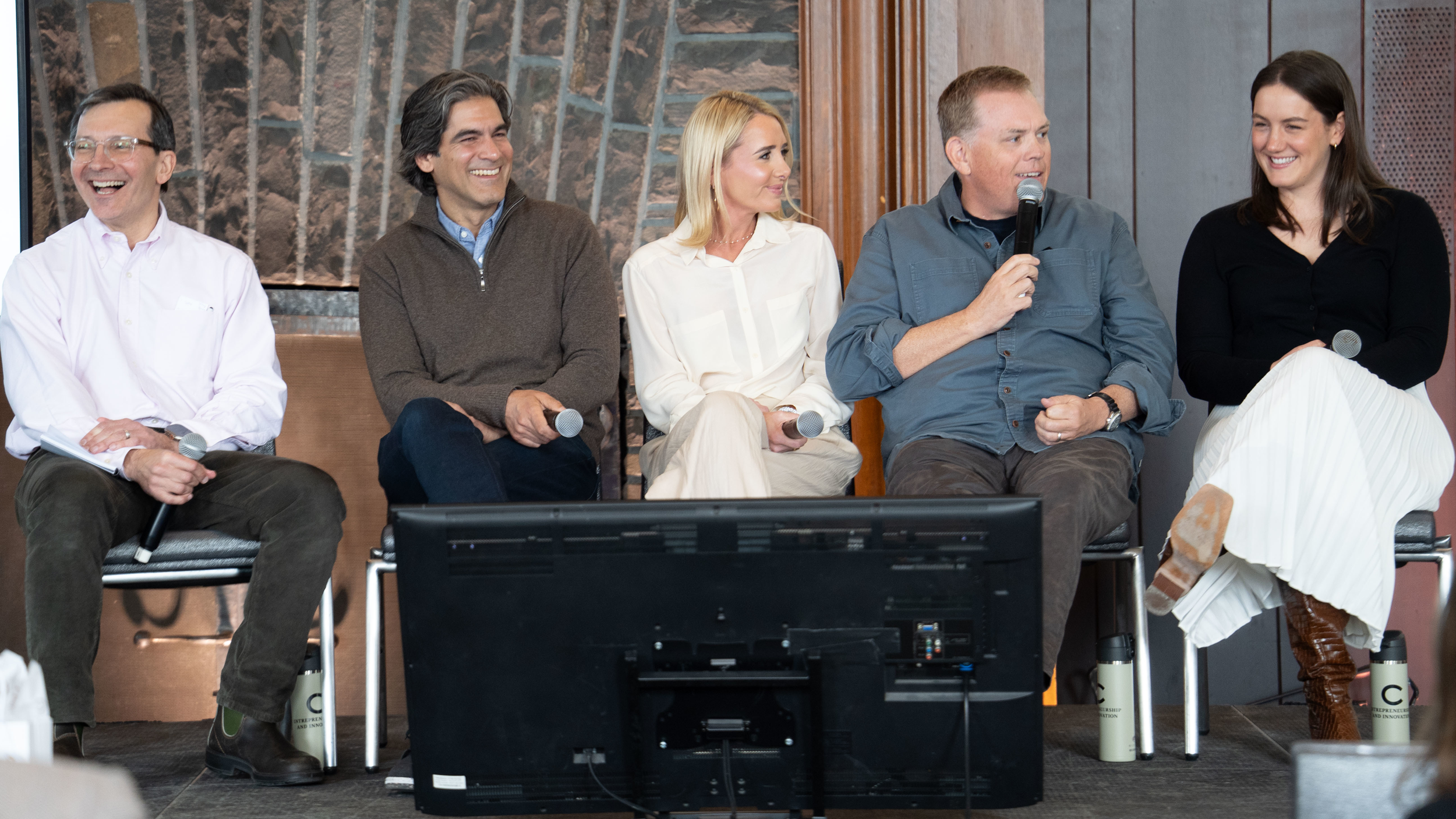 A panel of five people, three men and two women, are seated on a stage and laughing during a lively discussion. The individual second from the right speaks into a microphone while the others hold their own microphones and look on with engaged, joyful expressions.