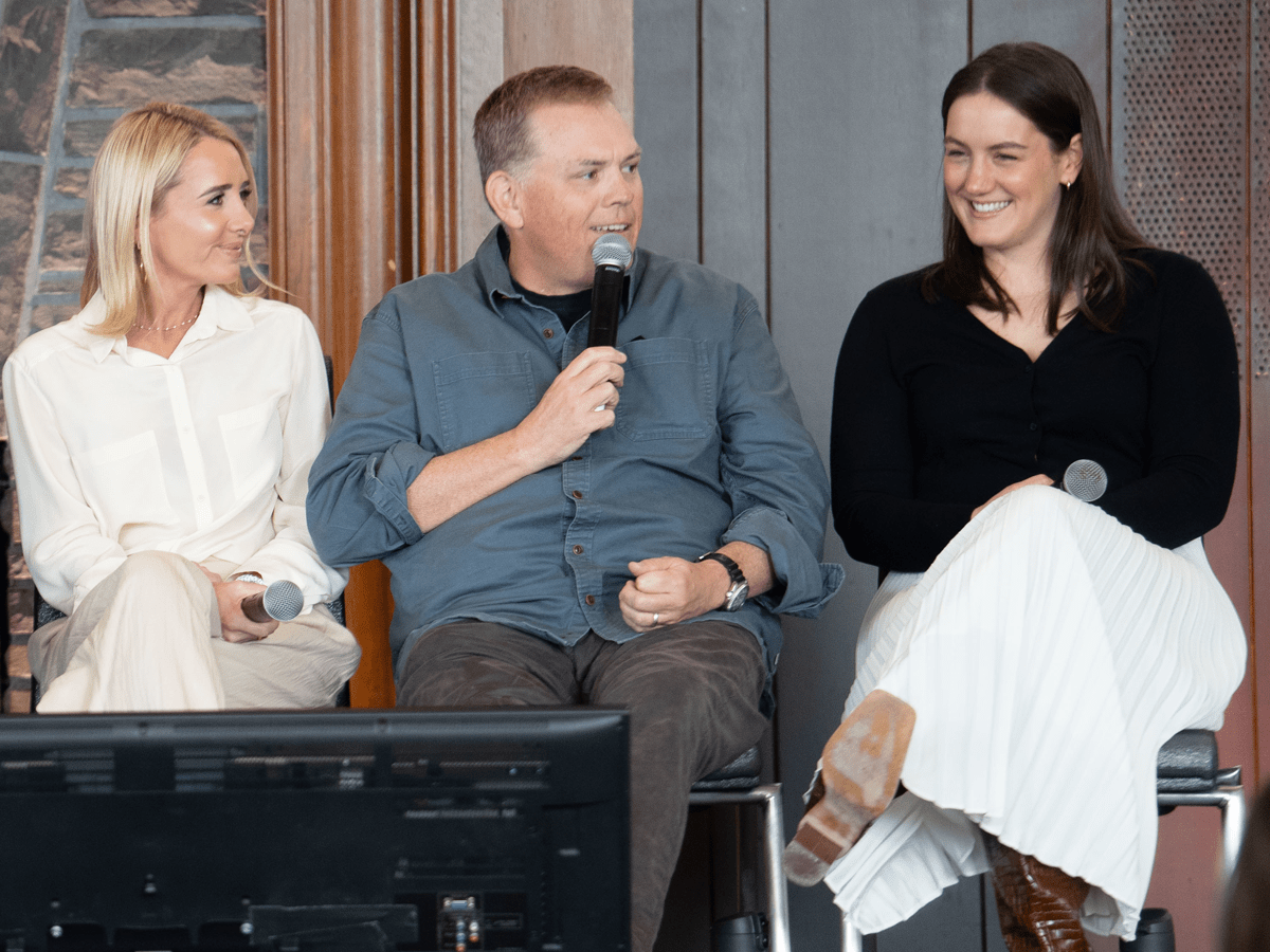 Three panelists, two women and one man, are seated together on a stage during a discussion or event. The man in the center speaks into a handheld microphone while the women on either side listen and smile, with a television monitor visible in the foreground.