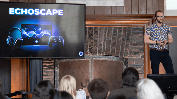 A presenter in a patterned short-sleeve shirt stands at the front of a room beside a large screen displaying the EchoScape logo and product imagery, while audience members watch from their seats.