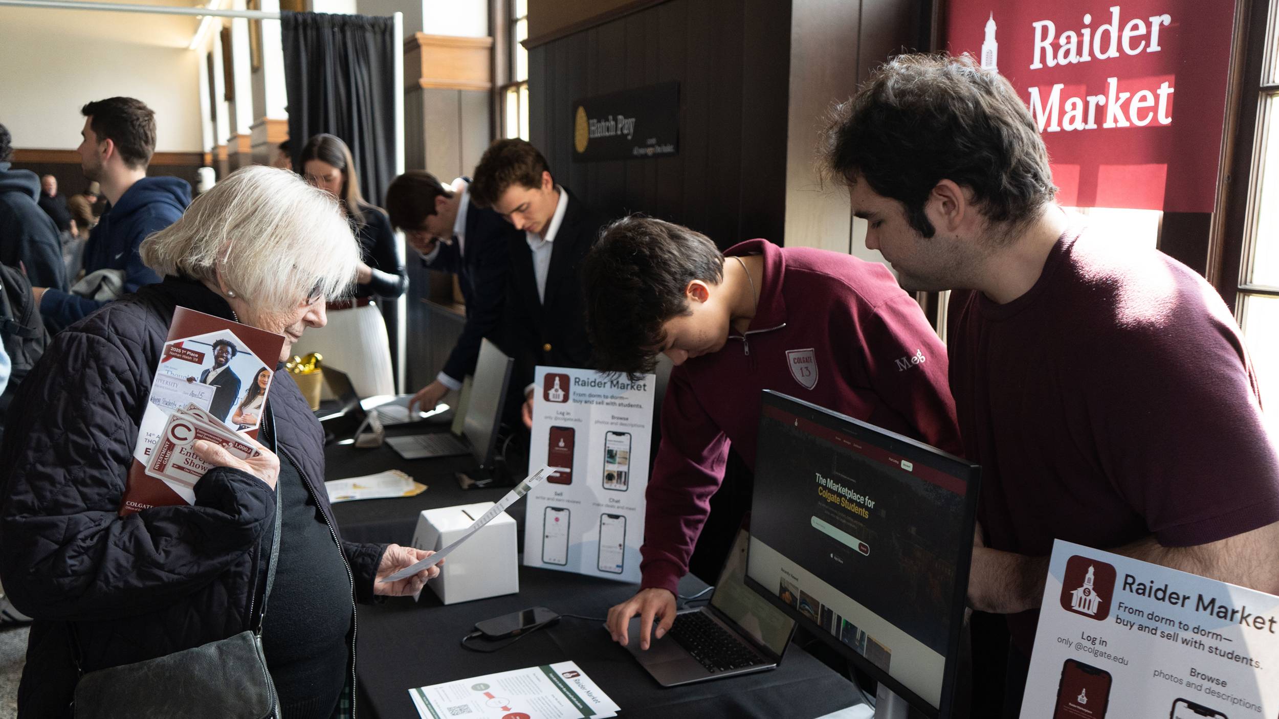 An older woman with white hair stands at a display table, reading a printed flyer, while two young men in maroon shirts demonstrate a website titled "The Marketplace for Colgate Students" on a laptop and monitor. The booth features signs for "Raider Market," a student marketplace platform, within a crowded indoor venue where other presentation tables are visible in the background.