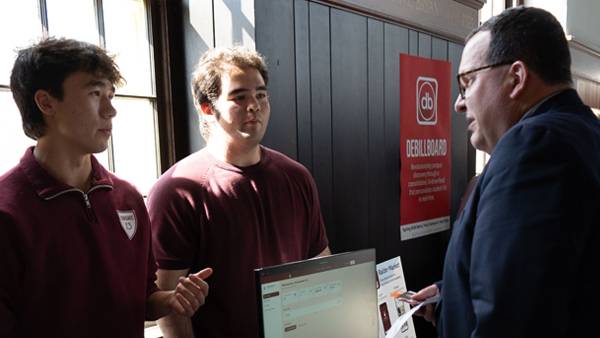 Two young men standing behind a table, dressed in maroon shirts, present a digital project on a computer monitor to an older man wearing glasses and a dark suit.