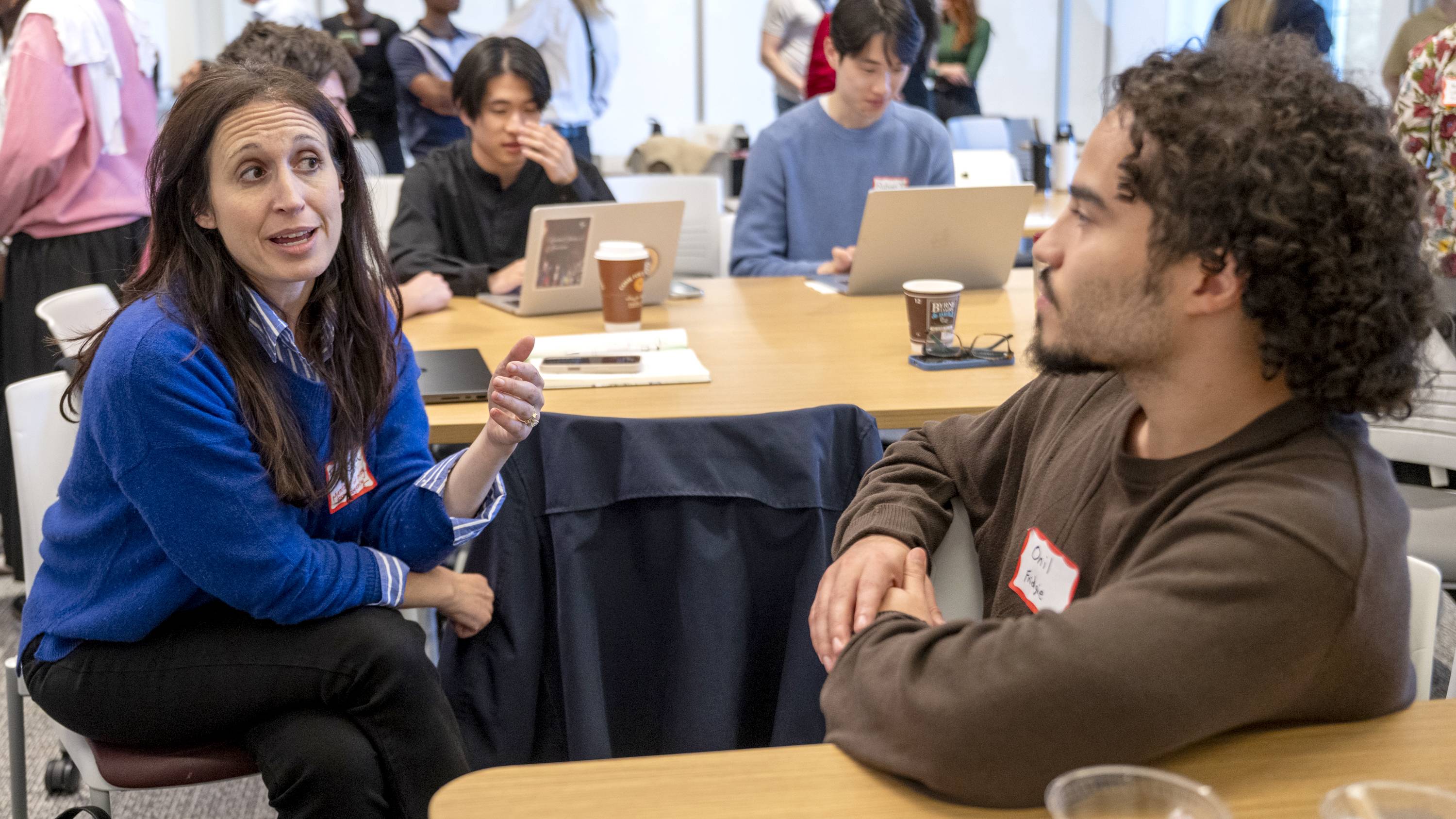 A woman with long dark hair, wearing a bright blue sweater over a striped button-down shirt, gestures with her hand while talking to a man with curly dark hair and a beard who is wearing a brown sweater. They are seated at a wooden table in a busy classroom or workshop setting where other individuals are working on laptops in the background.