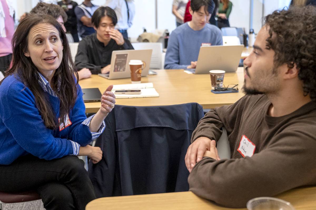 A woman with long dark hair, wearing a bright blue sweater over a striped button-down shirt, gestures with her hand while talking to a man with curly dark hair and a beard who is wearing a brown sweater. They are seated at a wooden table in a busy classroom or workshop setting where other individuals are working on laptops in the background.