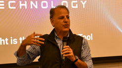 Mike Ellenbogen stands in front of a projection screen with the word technology on it, speaking into a microphone.