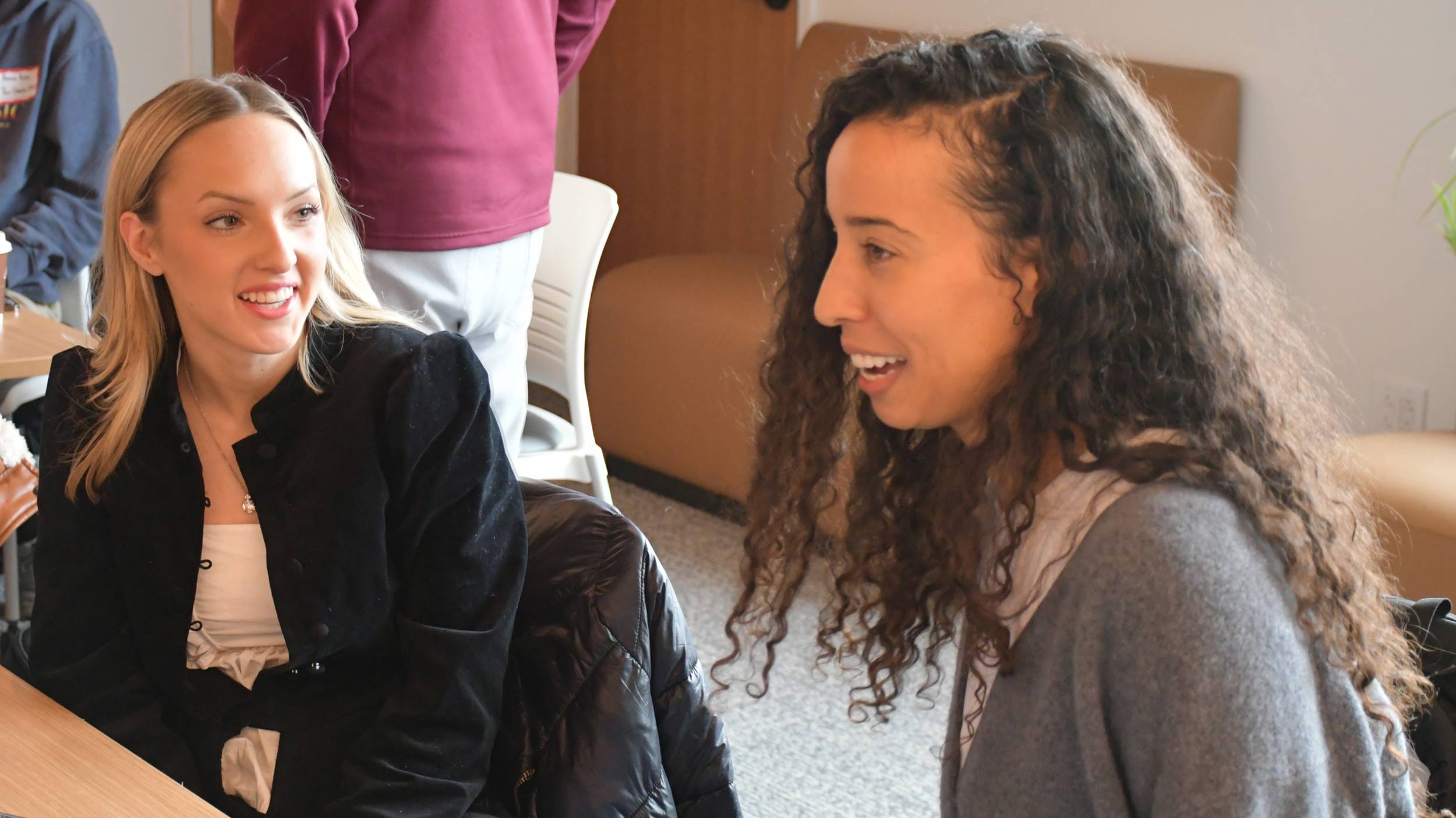 A candid, medium shot captures a young woman with long blonde hair seated at a table on the left and smiling as she converses with another person. To her right, a woman with long, curly dark hair is seen in profile, laughing during their interaction in a casual classroom setting.