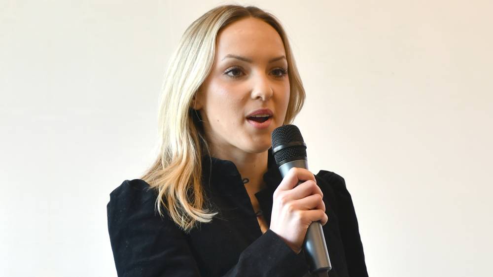 A young woman with long blonde hair is shown from the chest up as she speaks into a handheld microphone. She is wearing a high-collared black jacket and is positioned slightly off-center against a plain, light-colored background.