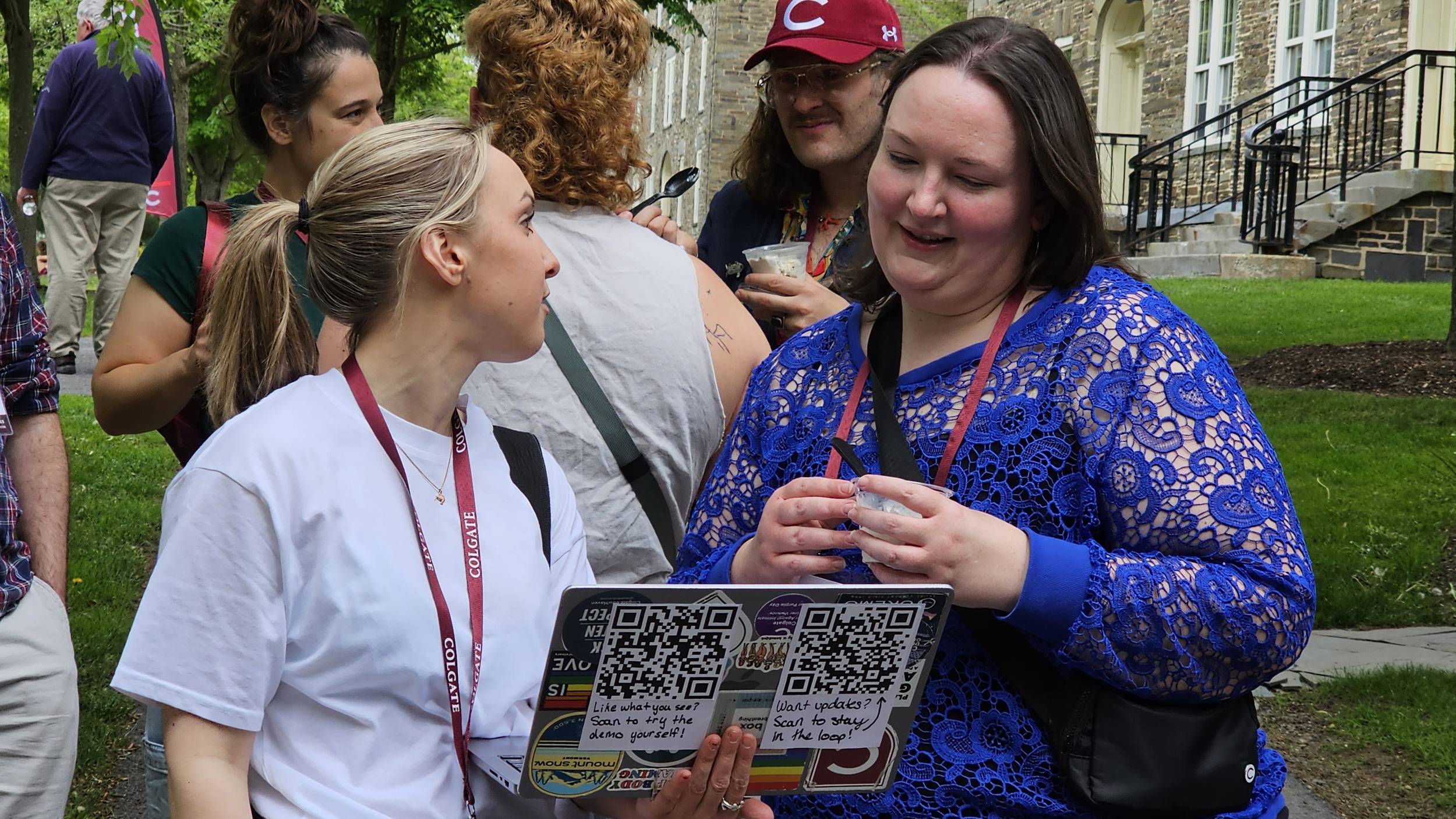 Two women stand together outdoors on a grassy campus lawn, with one in a white t-shirt holding open a sticker-covered laptop to show the other woman in a blue lace top. Both women are wearing maroon lanyards, and other event attendees are visible talking in the background near a stone building.