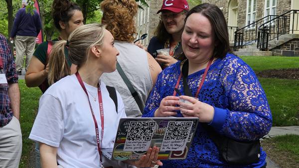 Two women stand together outdoors on a grassy campus lawn, with one in a white t-shirt holding open a sticker-covered laptop to show the other woman in a blue lace top. Both women are wearing maroon lanyards, and other event attendees are visible talking in the background near a stone building.