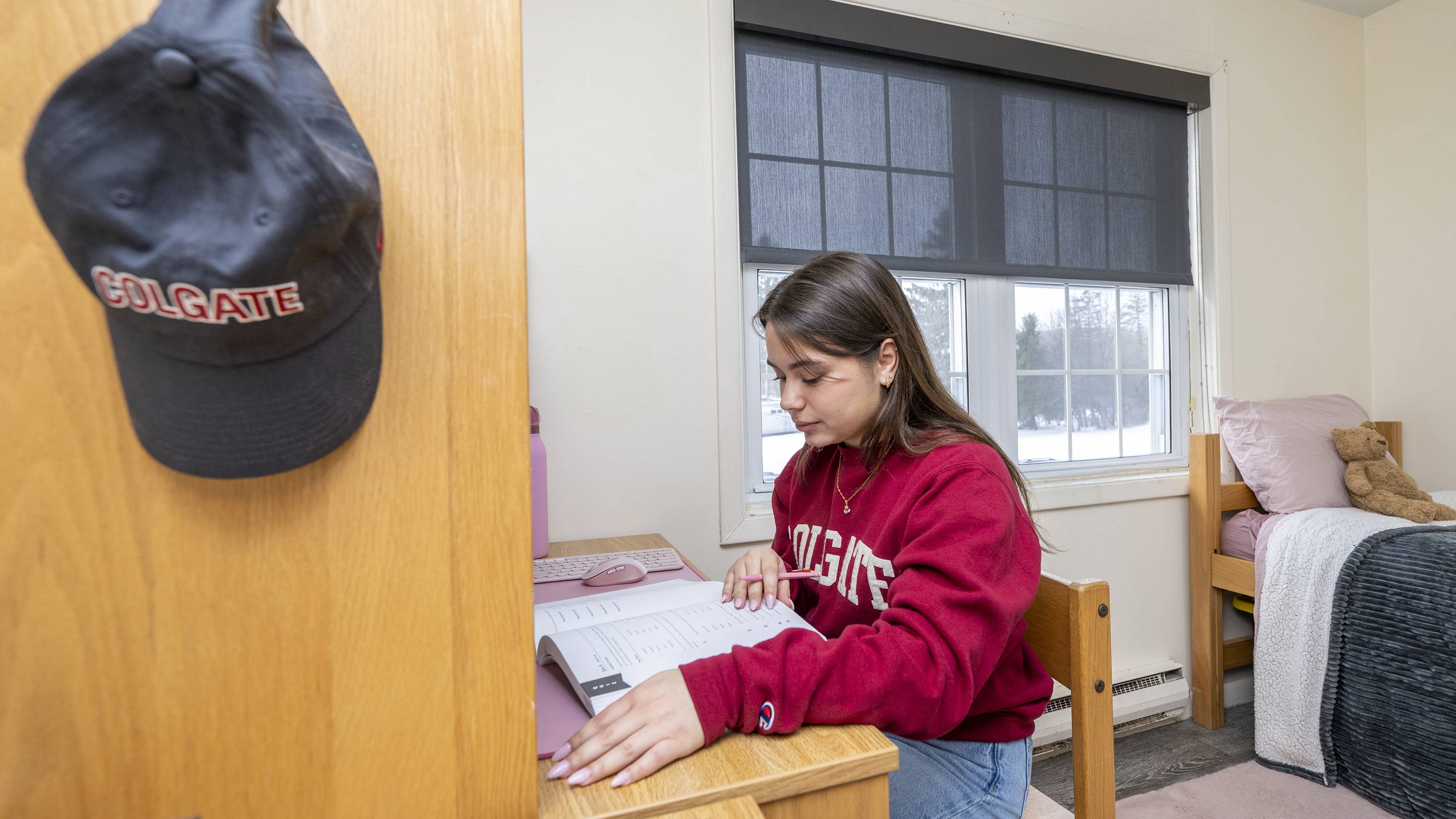 A student sits at a desk reading a book in a dorm room