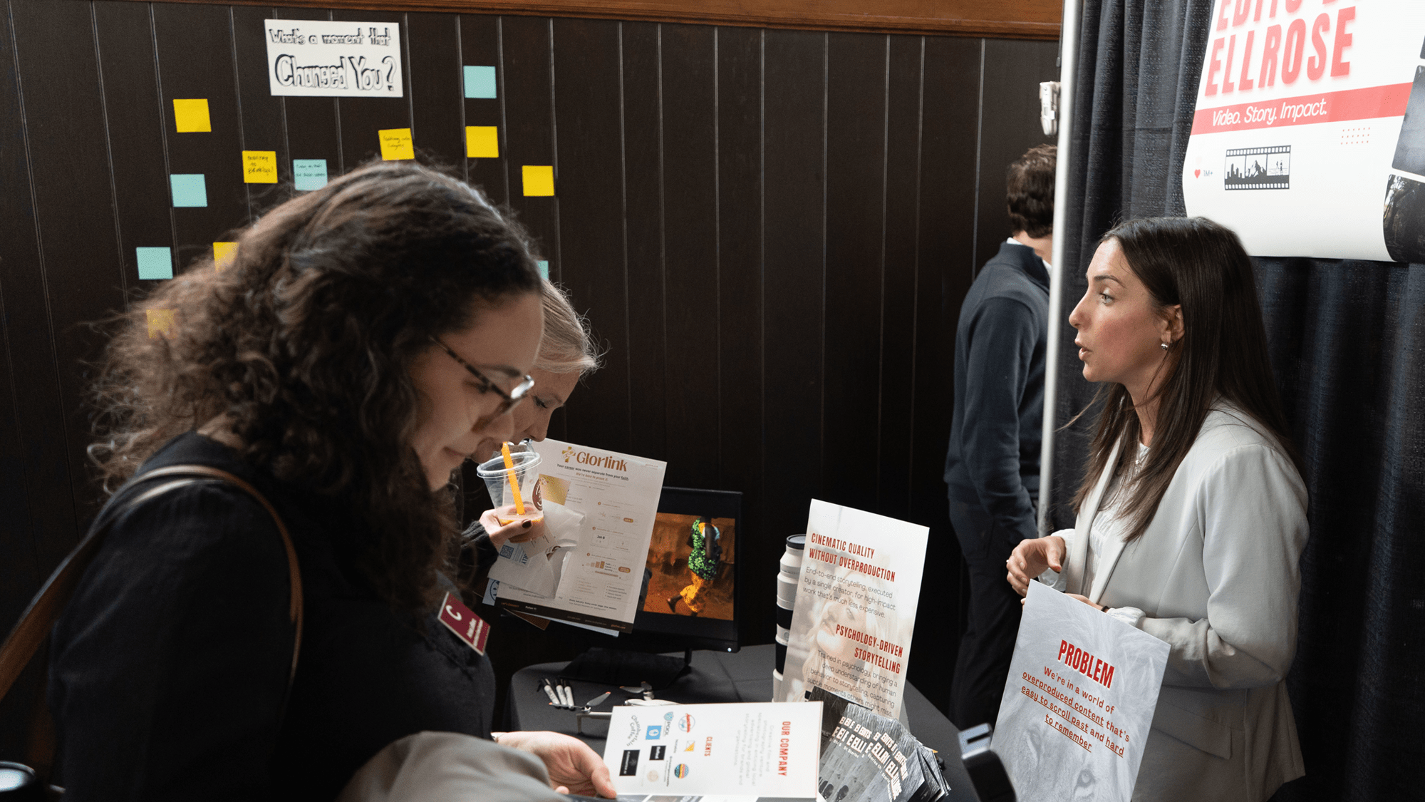 A woman in a white blazer stands behind a display table, speaking with two attendees who are looking at marketing brochures for "Ellrose." The booth features several signs with red and black text, while a dark wall in the background is decorated with colorful sticky notes and a sign asking, "What's a moment that changed you?"