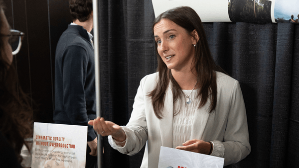 A woman with long brown hair, wearing a white blazer and necklace, stands at a promotional booth and gestures with her hand while speaking to an attendee. She is surrounded by black curtains and marketing signs, one of which features the heading "Cinematic Quality Without Overproduction."
