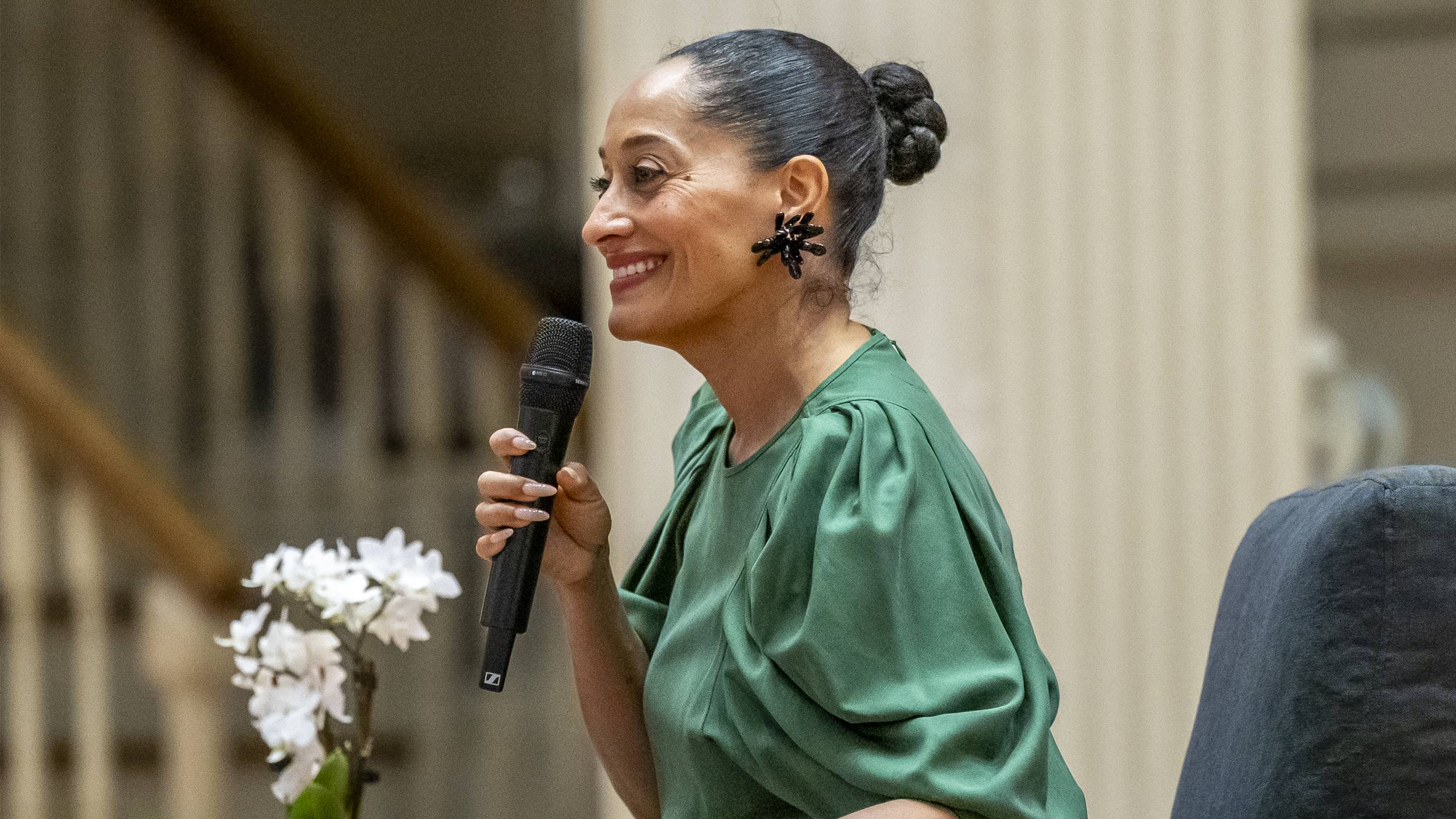 Tracee Ellis Ross is captured in profile, smiling warmly while holding a black microphone and appearing to speak at an event. She is wearing a vibrant green blouse with pleated sleeves, a bold black sculptural earring, and her hair is styled in a sleek high bun.