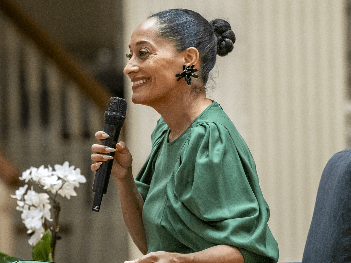 Tracee Ellis Ross is captured in profile, smiling warmly while holding a black microphone and appearing to speak at an event. She is wearing a vibrant green blouse with pleated sleeves, a bold black sculptural earring, and her hair is styled in a sleek high bun.