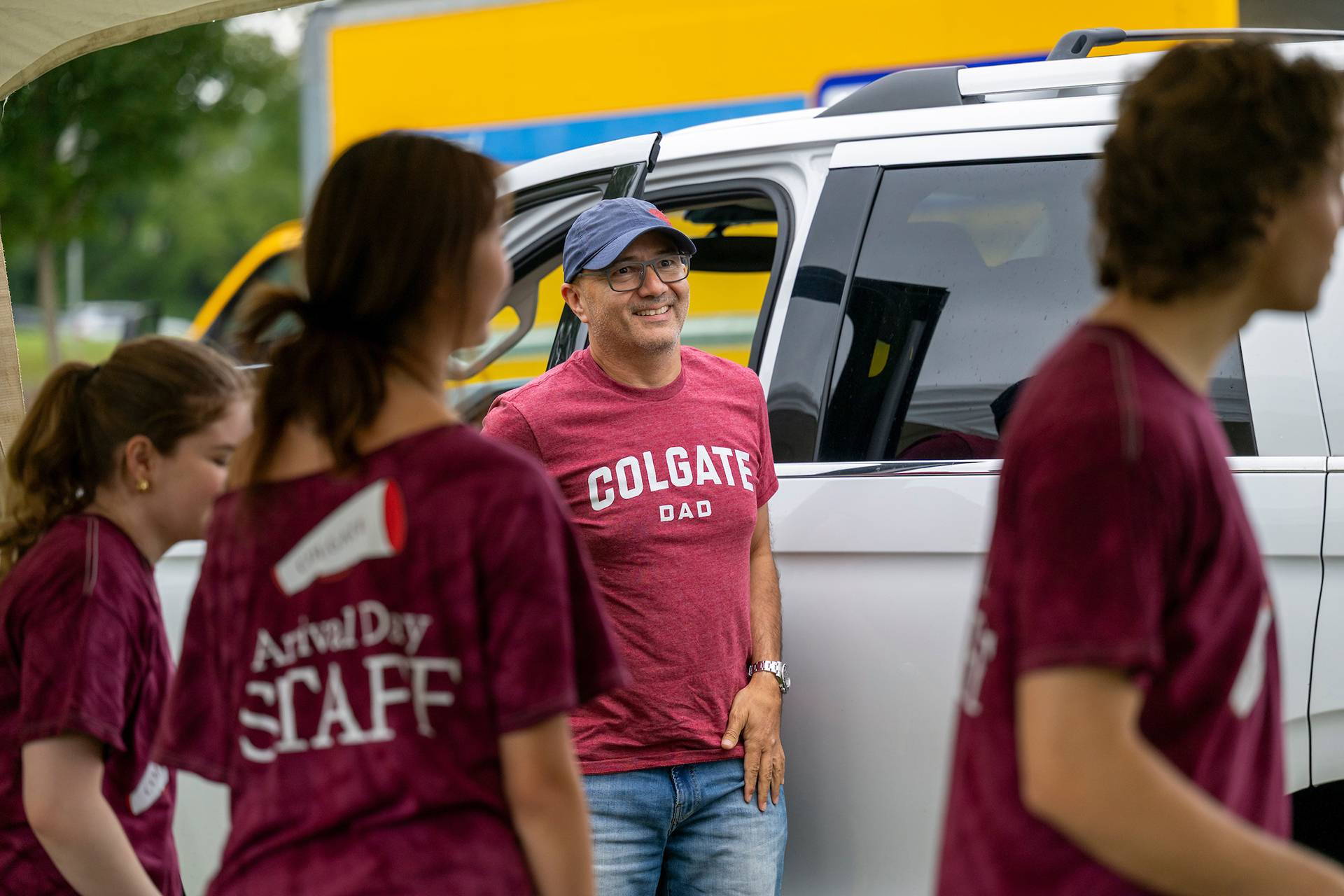 A Colgate parent wears a "Colgate Dad" t-shirt and smiles at student staff who are helping move incoming students belongings into dorm rooms.